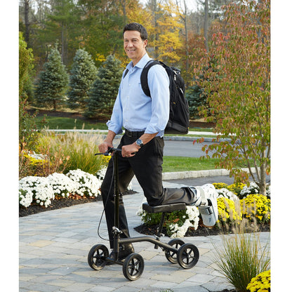 Man with a knee scooter walking outdoors on a paved path with trees and flowers in the background.