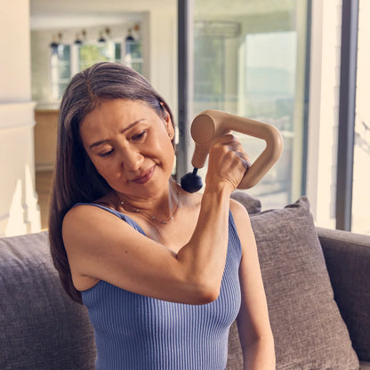 Woman using the sand-colored Theragun Relief massage gun on her shoulder for muscle recovery, tension relief, and relaxation.