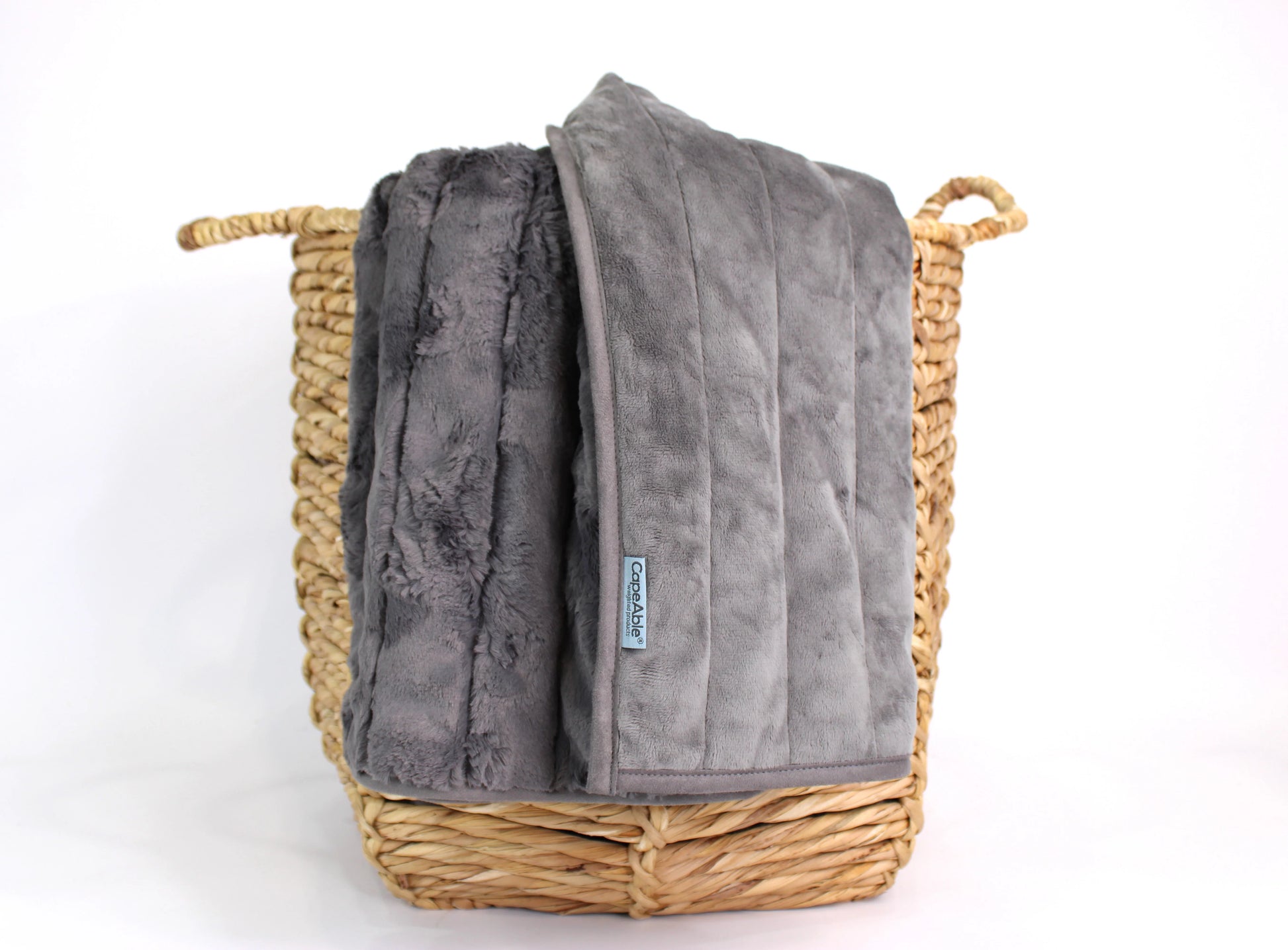 CapeAble weighted blanket in mid-century ash draped over a woven basket showing ribbed plush exterior and quilted gray interior lining, on white background