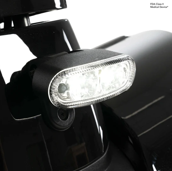 A close-up of a black medical device light with an illuminated clear lens, mounted on a black vehicle or equipment surface against a white background. Small text in the top right reads 'FDA Class II Medical Device'.