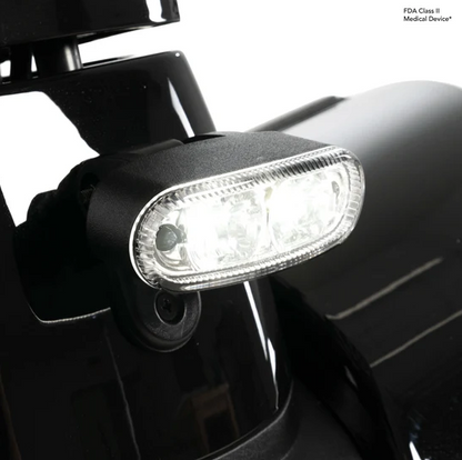 A close-up of a black medical device light with an illuminated clear lens, mounted on a black vehicle or equipment surface against a white background. Small text in the top right reads 'FDA Class II Medical Device'.
