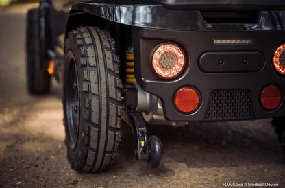 Close-up of a black medical scooter wheel with a small black wheeled medical device hanging from a hitch, on a dirt road. The vehicle has circular amber and red lights, and a yellow coil spring is visible behind the tire.